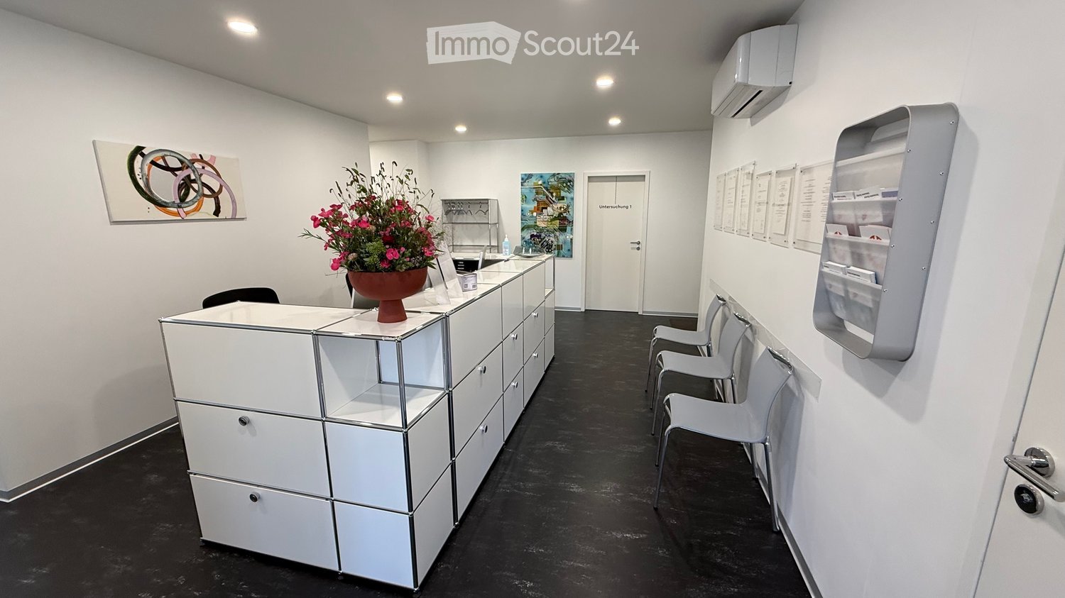 Modern reception area with white walls, black flooring, reception desk, chairs, flower vase, and framed artwork.