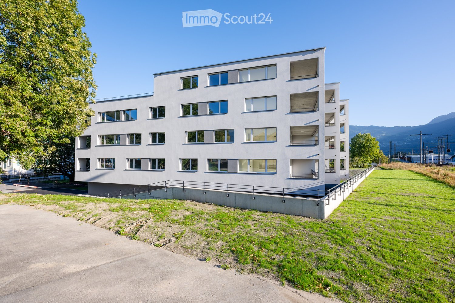 A white building with many windows and balconies, multiple floors, situated in a landscape with greenery and grass.
