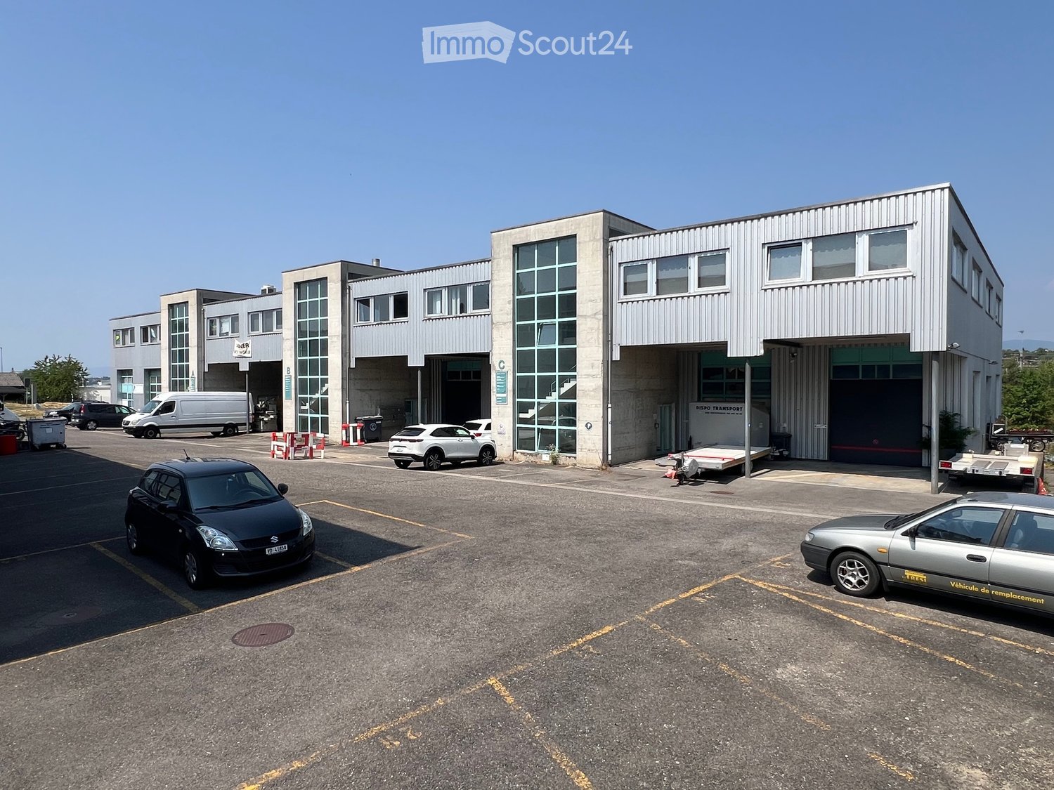 Modern industrial building with large garage doors, multiple windows, and a spacious parking lot with several cars.