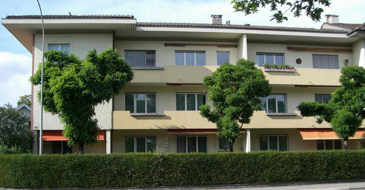 A three-story residential building with white and beige exterior, multiple windows, balconies, and surrounding greenery.