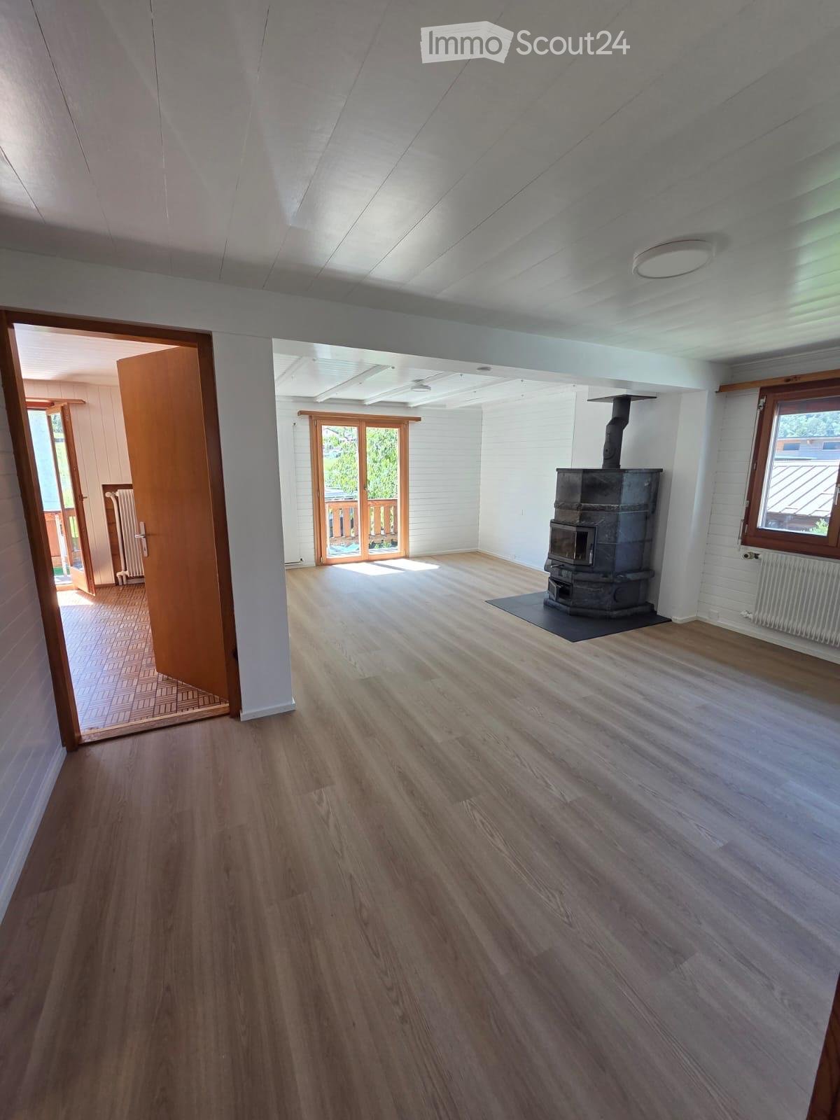 Empty room with wooden flooring, white walls, and a wood-burning stove. Natural light streams in from windows and an open doorway leading to another room.