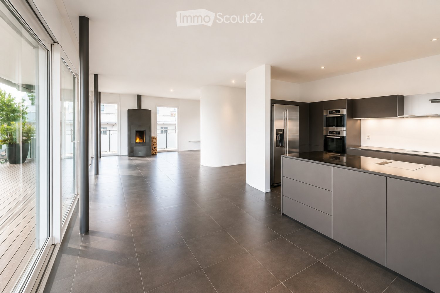 Modern kitchen with stainless steel appliances, dark grey cabinets, and a central island. Features a fireplace and large windows with a view of greenery.