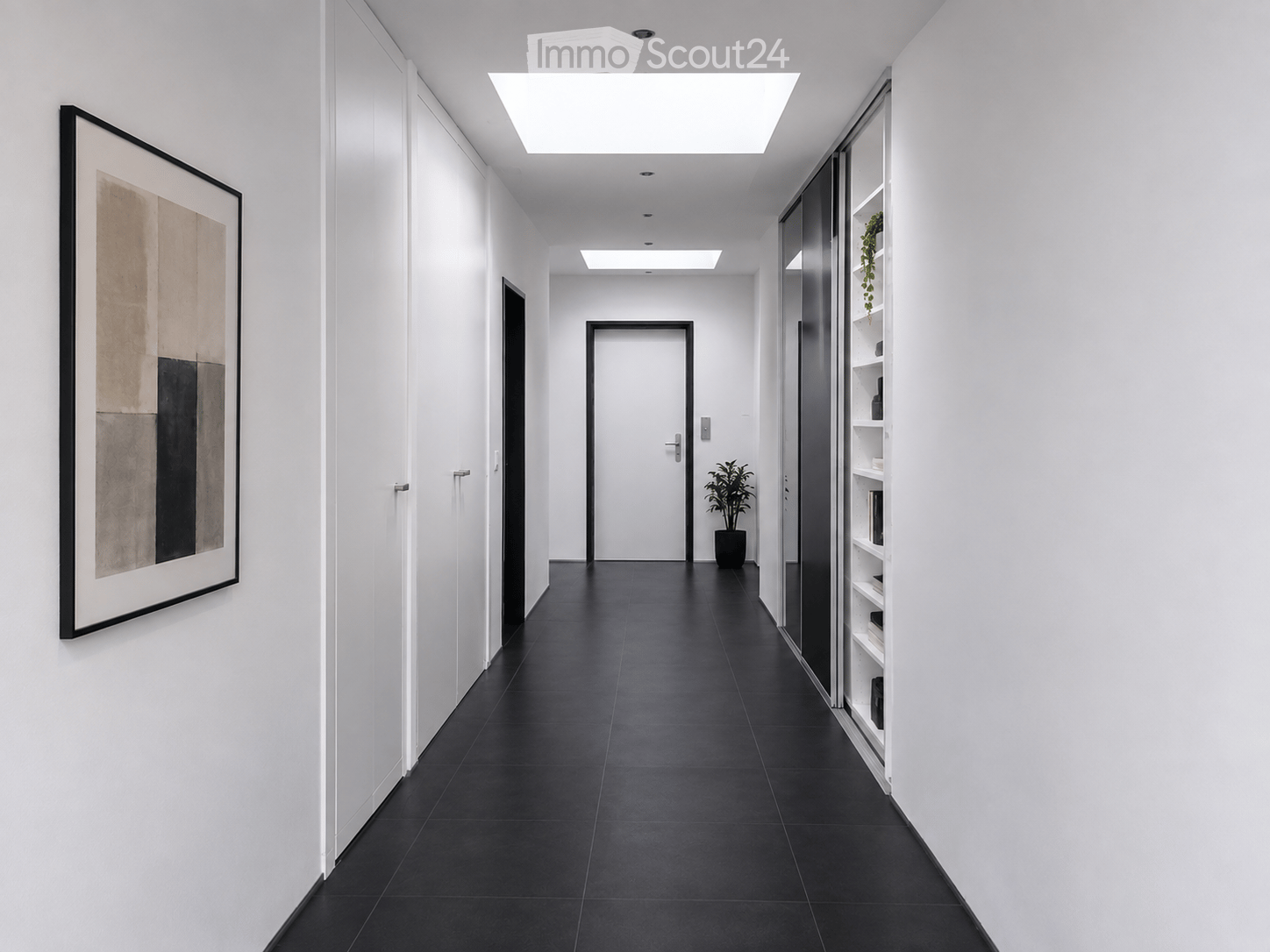 Modern hallway with white walls, black tiled floor, recessed ceiling lights, and a large framed artwork on the left wall. A potted plant sits near the door at the end of the corridor.