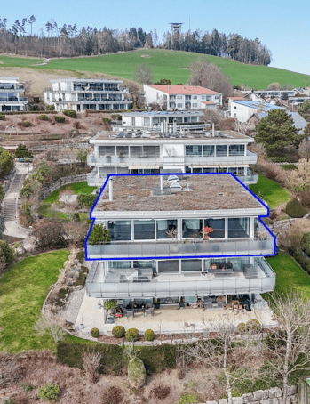Aerial view of a large, modern multi-story house with a flat roof and extensive windows, surrounded by a landscaped garden and other residential buildings in the background.