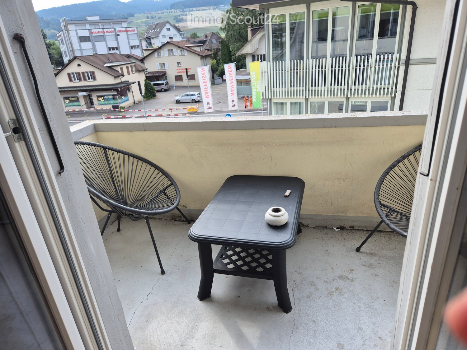Balcony with a small black table and two black chairs, overlooking a town with buildings and a scenic landscape.