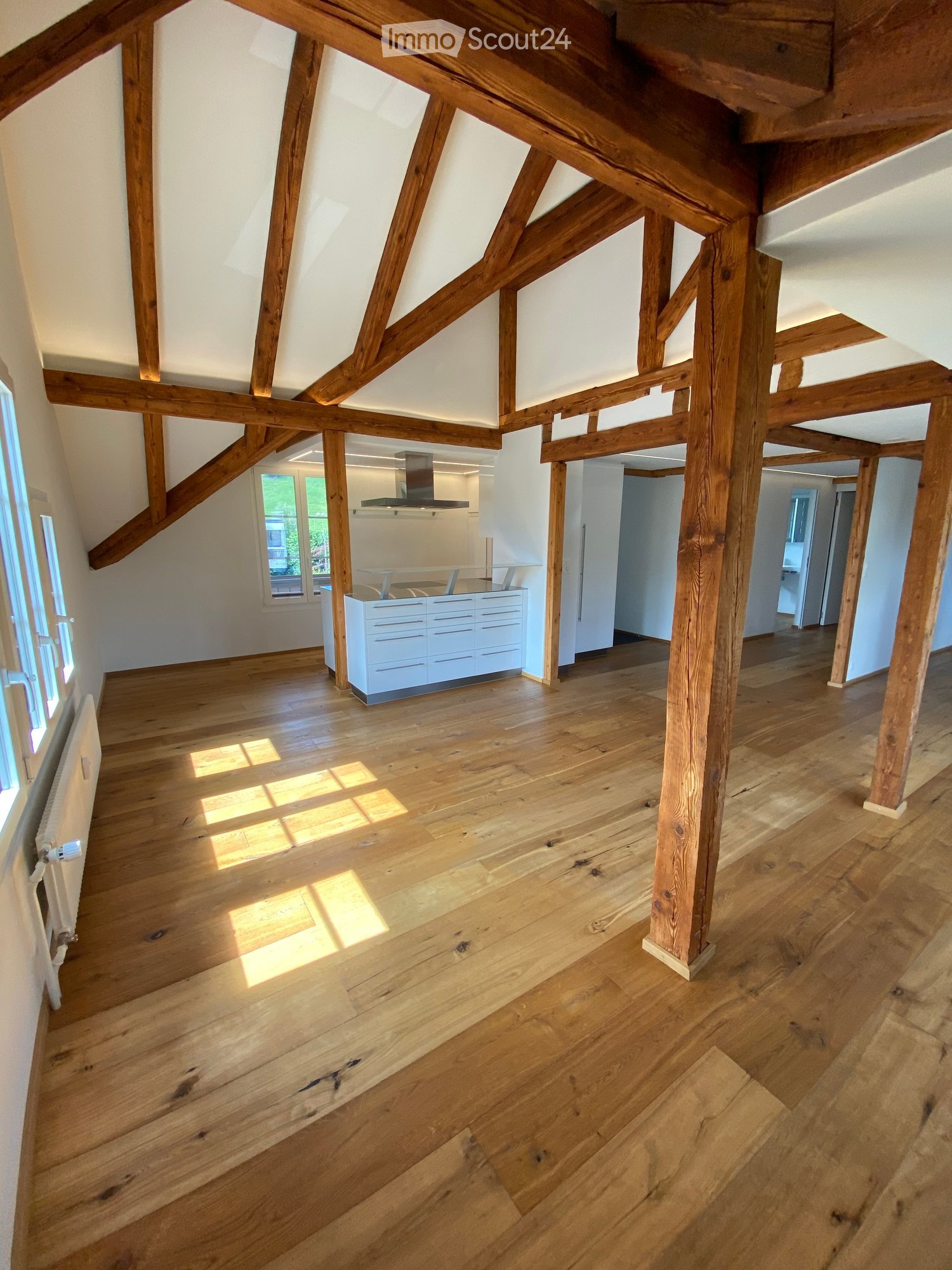 Open, high-ceilinged room with exposed wooden beams and polished wooden flooring. Natural light streams in from large windows, illuminating the space.