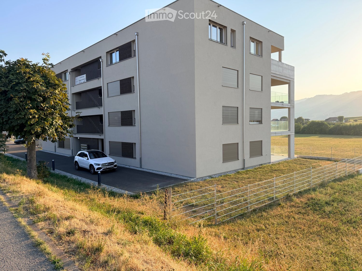 Modern multi-story apartment building with large windows, balconies, and a parking area.