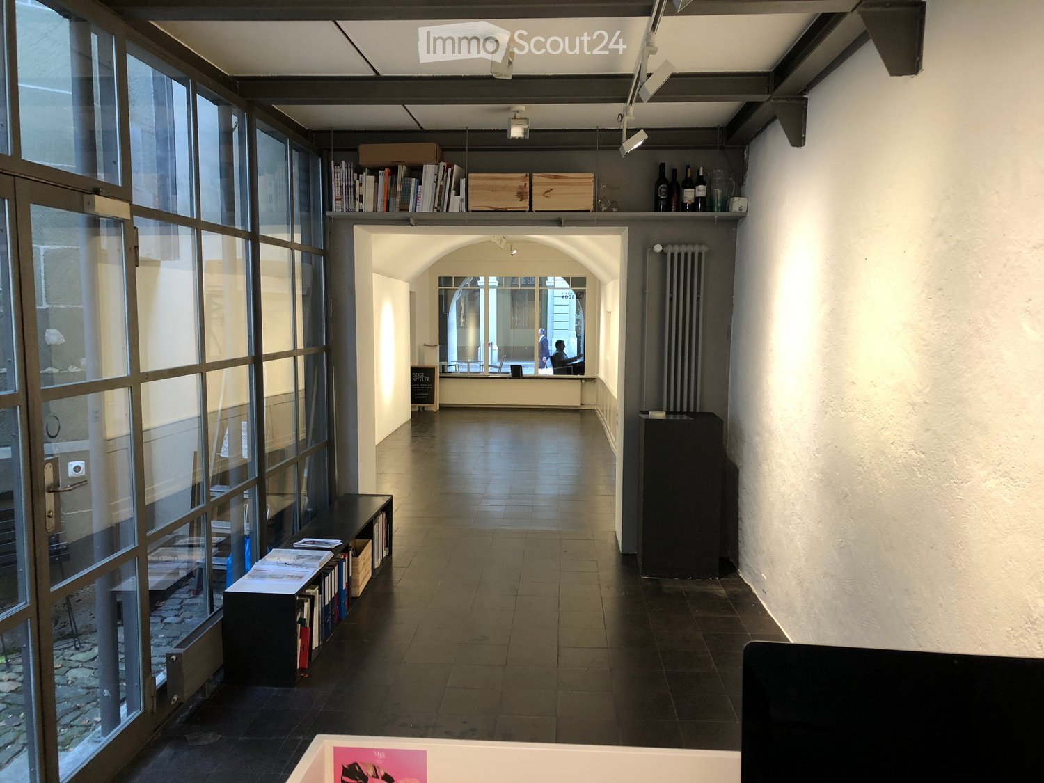A modern hallway with large glass windows on one side, wooden shelves holding books and boxes, and a black cabinet on the right. The floor is tiled, and the ceiling has exposed beams and track lighting.