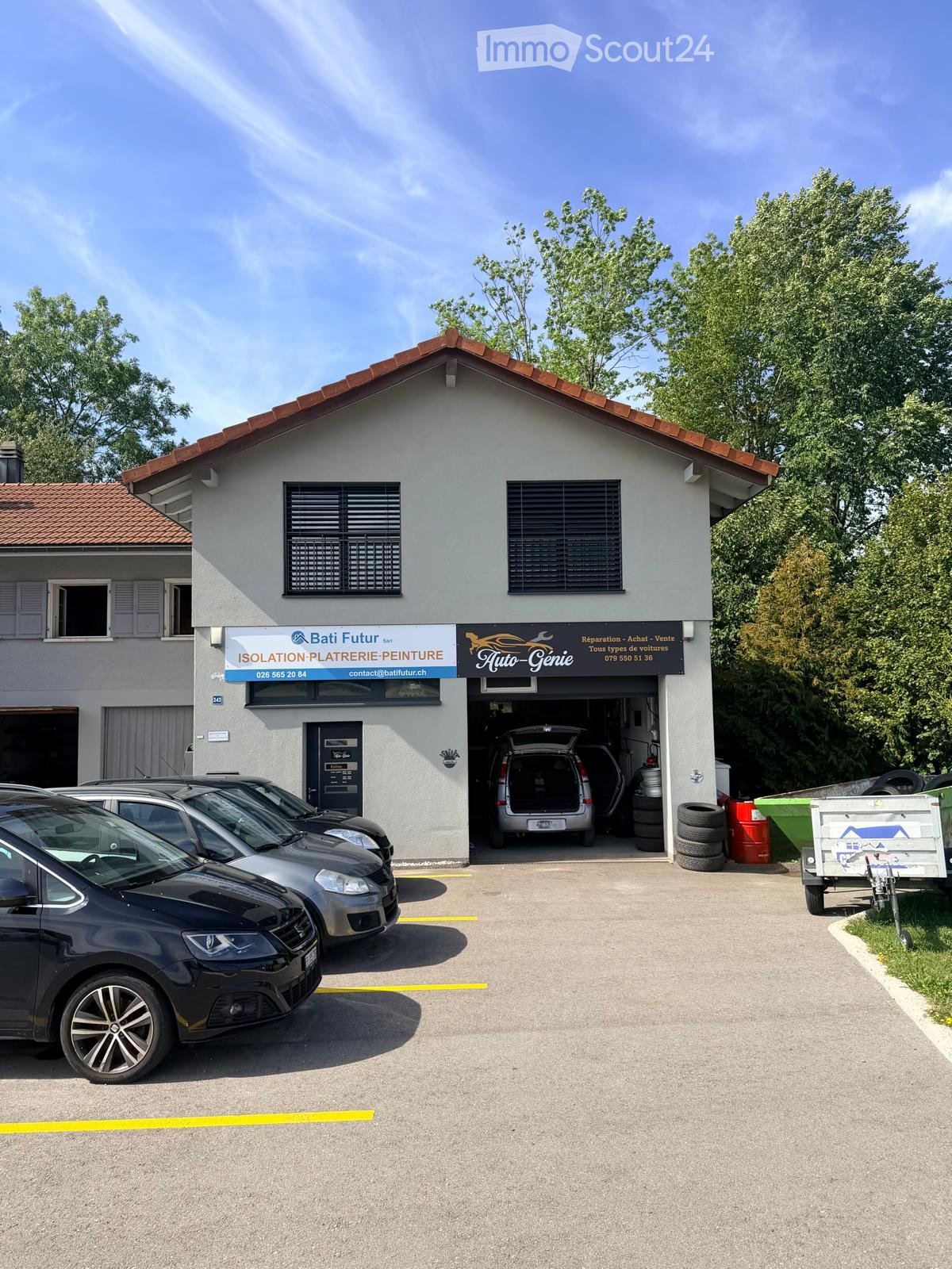 A two-story building with a garage, featuring a sign for "Bati Futur" and "Auto-Genie" services. Several cars are parked in front, and the garage door is open, revealing a vehicle inside.