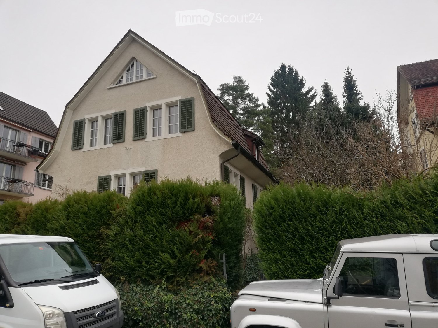 Two-story house with beige siding, green shutters, and a triangular roof window. The house is surrounded by tall evergreen hedges and trees, with two parked vans in the foreground.