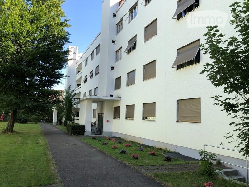 Modern multi-story building with white facade, multiple windows, and a small garden area with flowers and a tree.