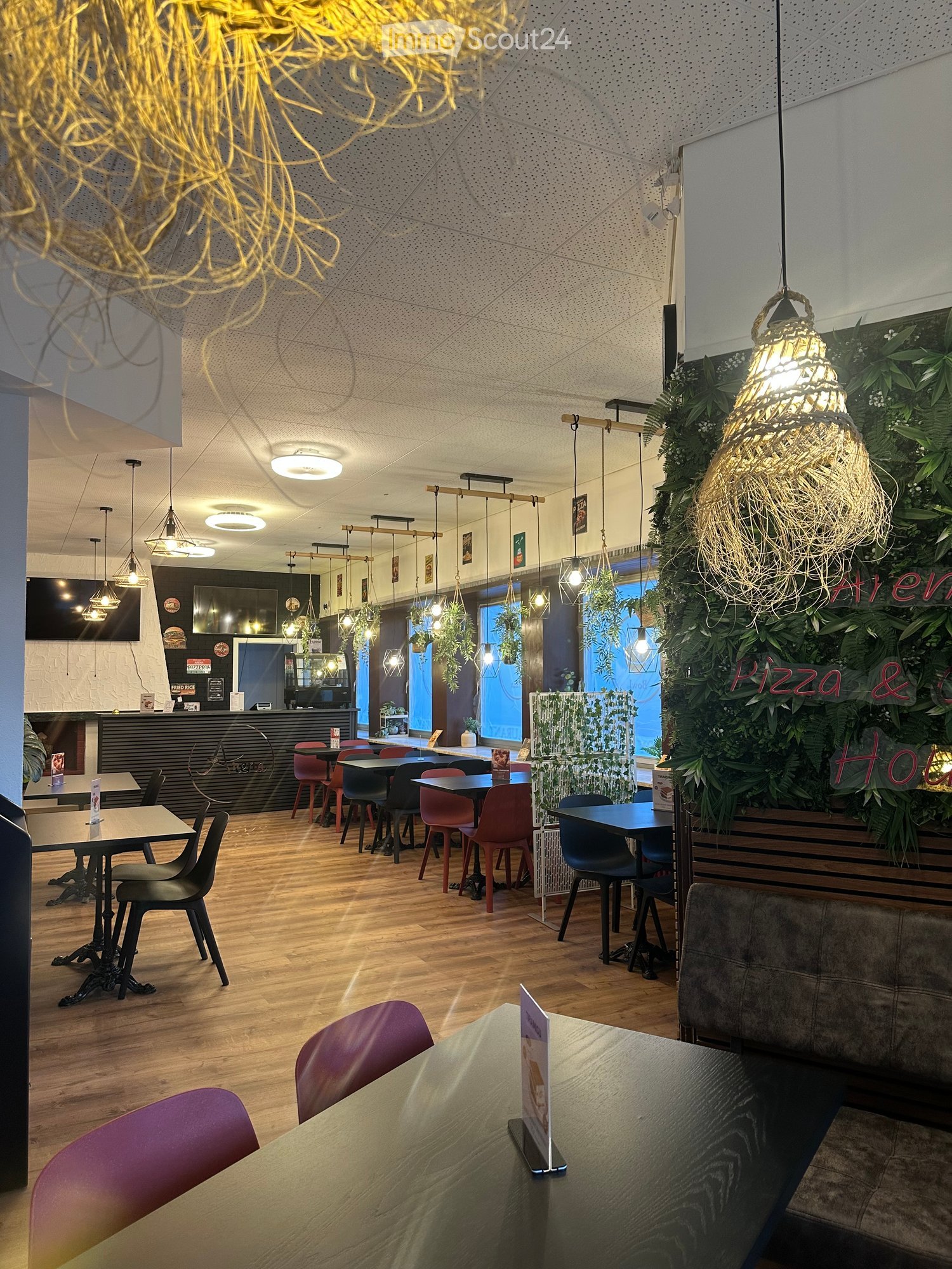 Modern restaurant interior with wooden flooring, pendant lights, tables, chairs, and a green wall with hanging plants.