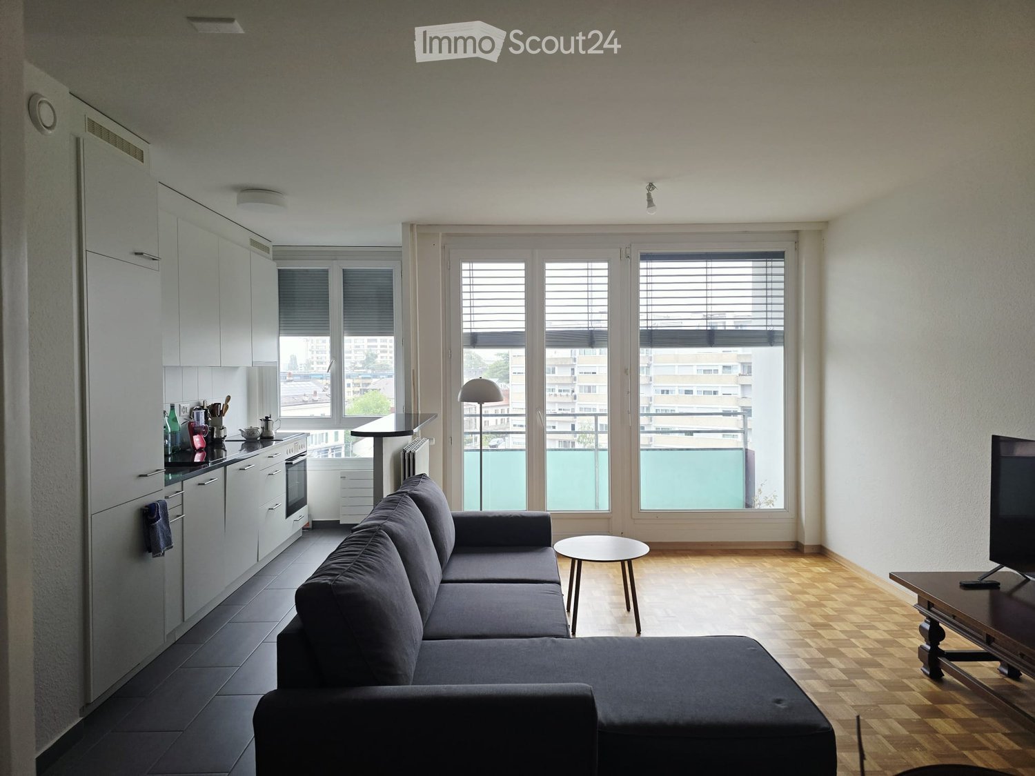Modern living room with a dark grey sectional sofa, coffee table, TV, and a small kitchen area with cabinets, oven, and fridge. Large windows with blinds overlook a balcony.