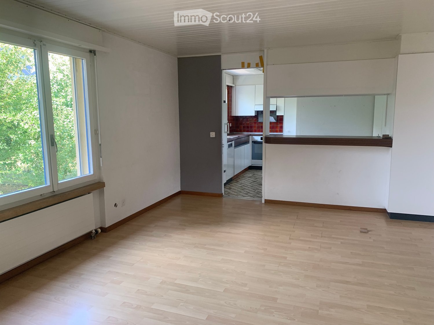 Empty room with wooden flooring, white walls, large window with view of greenery, and an open doorway leading to a small kitchen area.