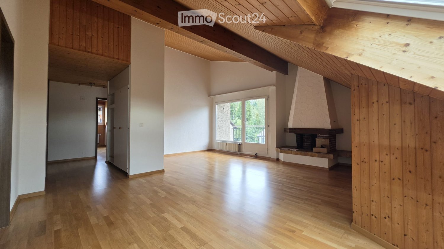 Empty room with wooden flooring, exposed wooden beams on the ceiling, white walls, and a fireplace with a bench. There is a window with a view of greenery and a door leading to another room.