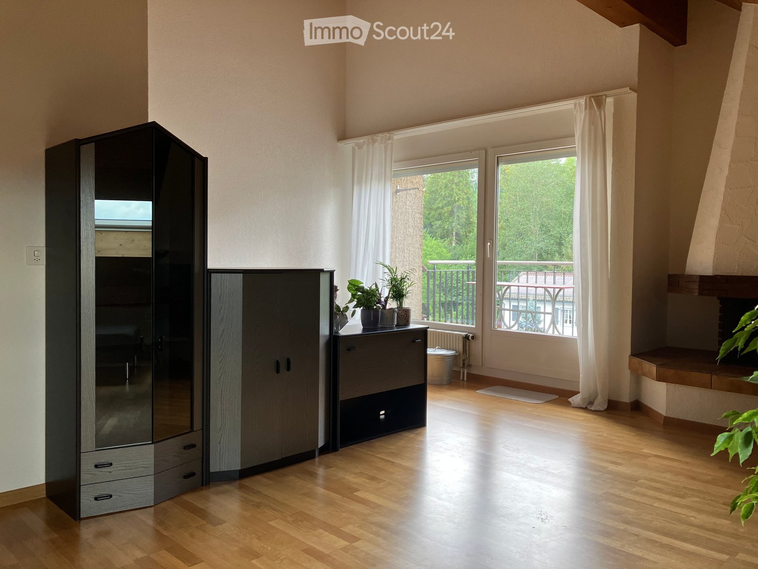 Bright room with wooden flooring, large windows leading to a balcony, modern wardrobe, small cabinet with potted plants on top, and a trash can near the window.