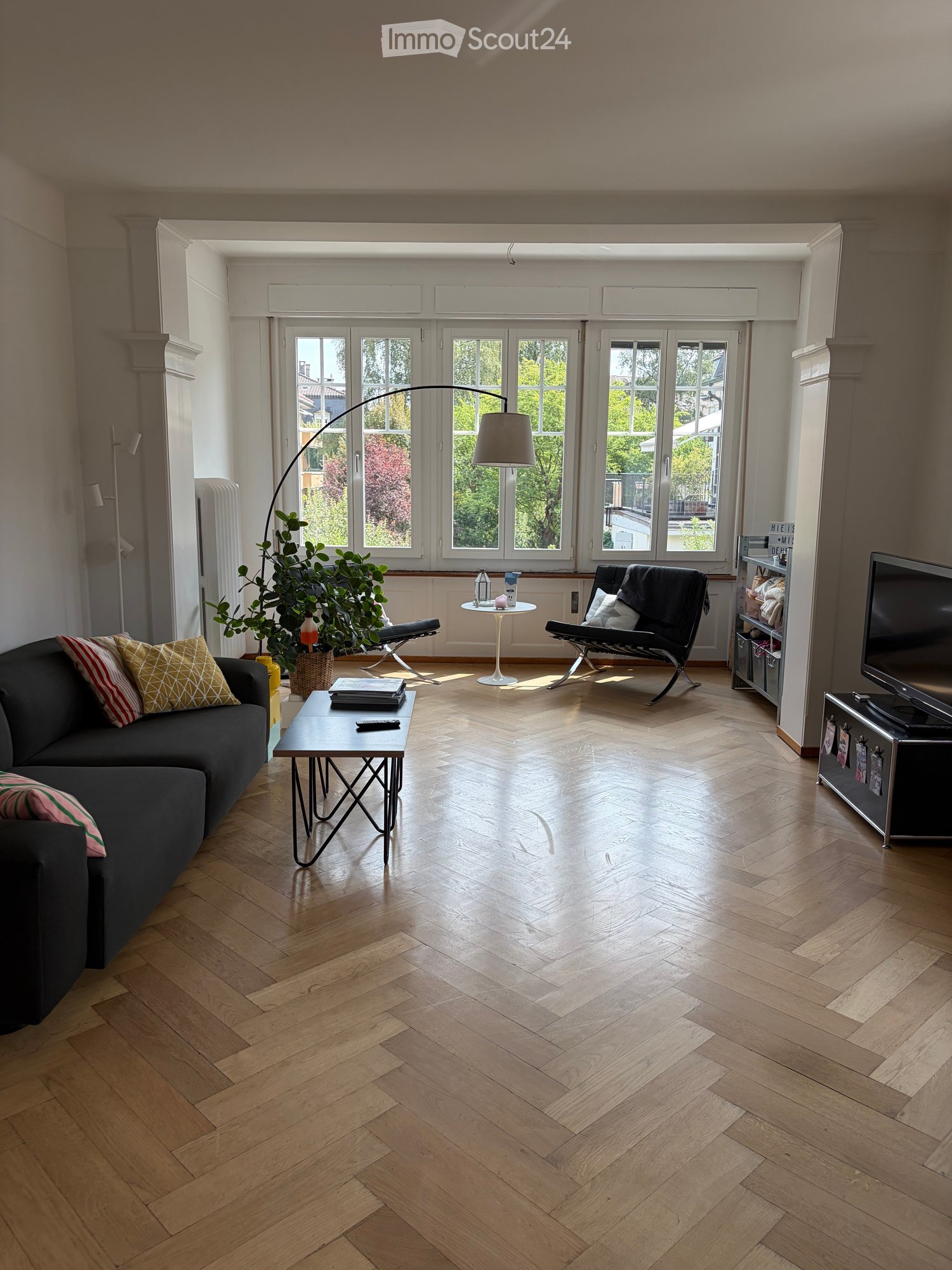 Modern living room with wooden herringbone flooring, large windows, a dark grey sofa with patterned cushions, a black coffee table, a white side table, a floor lamp, a TV stand, and a potted plant.