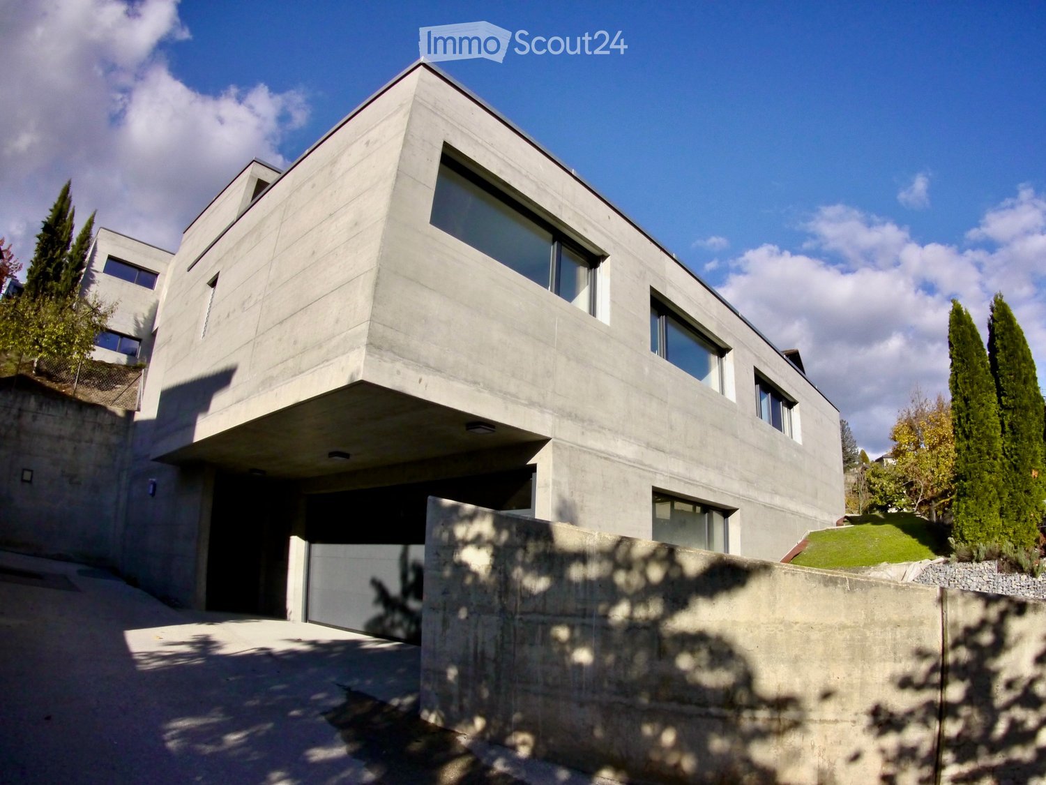 Modern two-story concrete house with large windows, raised ground floor, and surrounding greenery.
