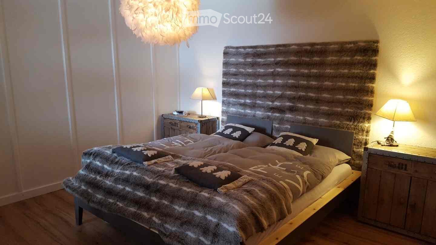 Cozy bedroom with a large bed, fur throw, decorative pillows, nightstands, lamps, and a textured headboard.