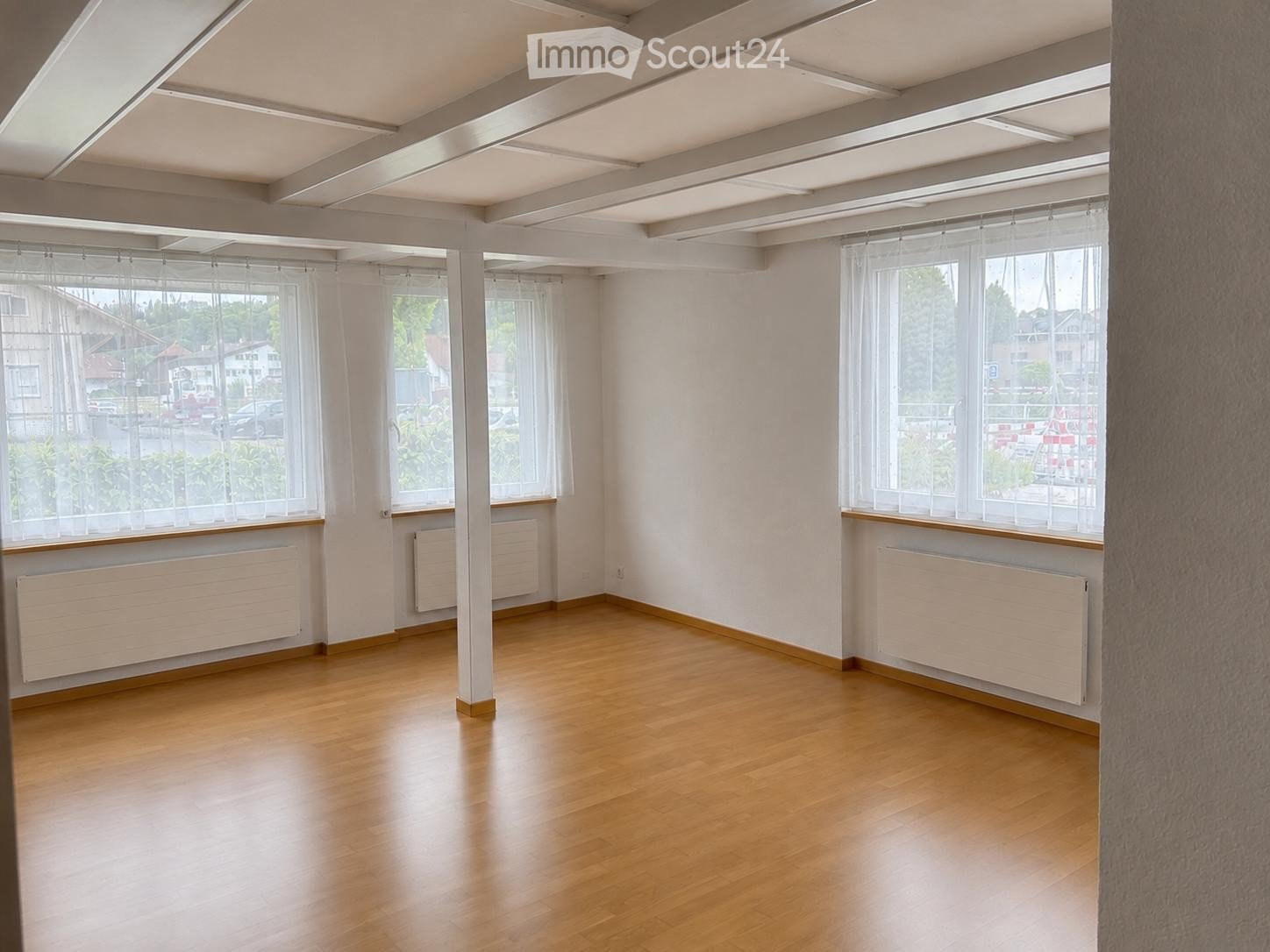 Empty room with wooden flooring, white walls, large windows with sheer curtains, radiators, and exposed ceiling beams.