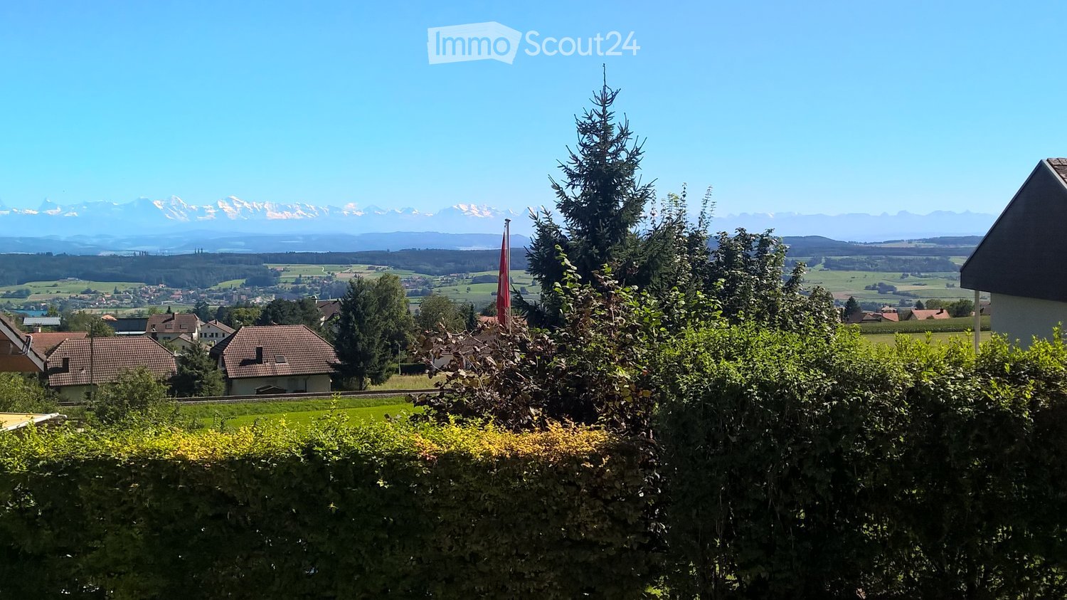 A scenic outdoor view with a hedge in the foreground, houses and trees in the middle ground, and a panoramic mountain range in the background under a clear blue sky.