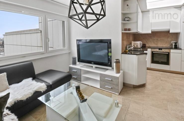 Modern, open-plan living and dining area with a black leather sofa, glass coffee table, flat-screen TV on a white stand, and a compact white kitchen with cabinets, sink, and oven. Large windows offer a city view.