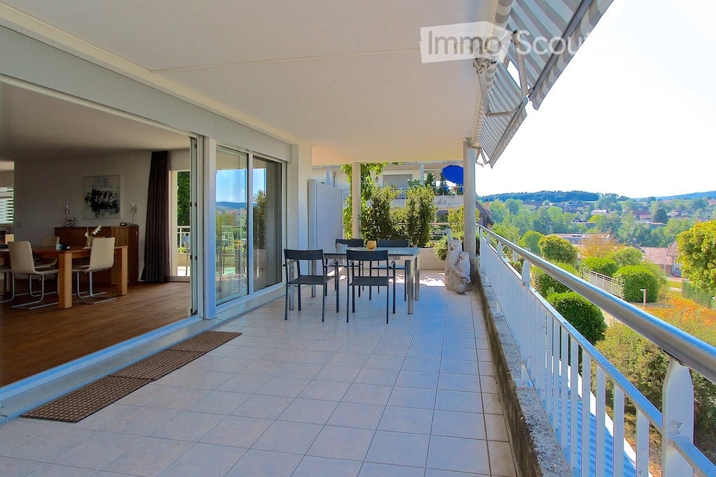 Spacious balcony with tiled floor, glass sliding doors leading to a dining area inside, outdoor dining set, and scenic view of greenery and hills.