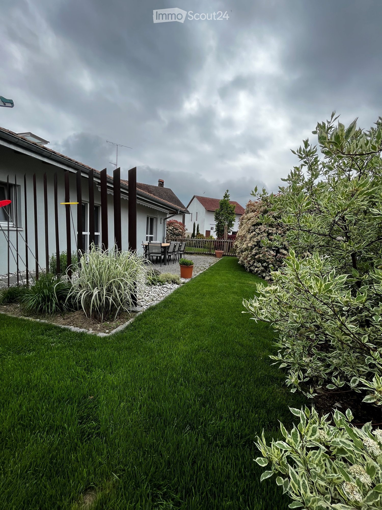 Well-maintained garden with a lawn, various plants, and a patio area with a table and chairs near the house.