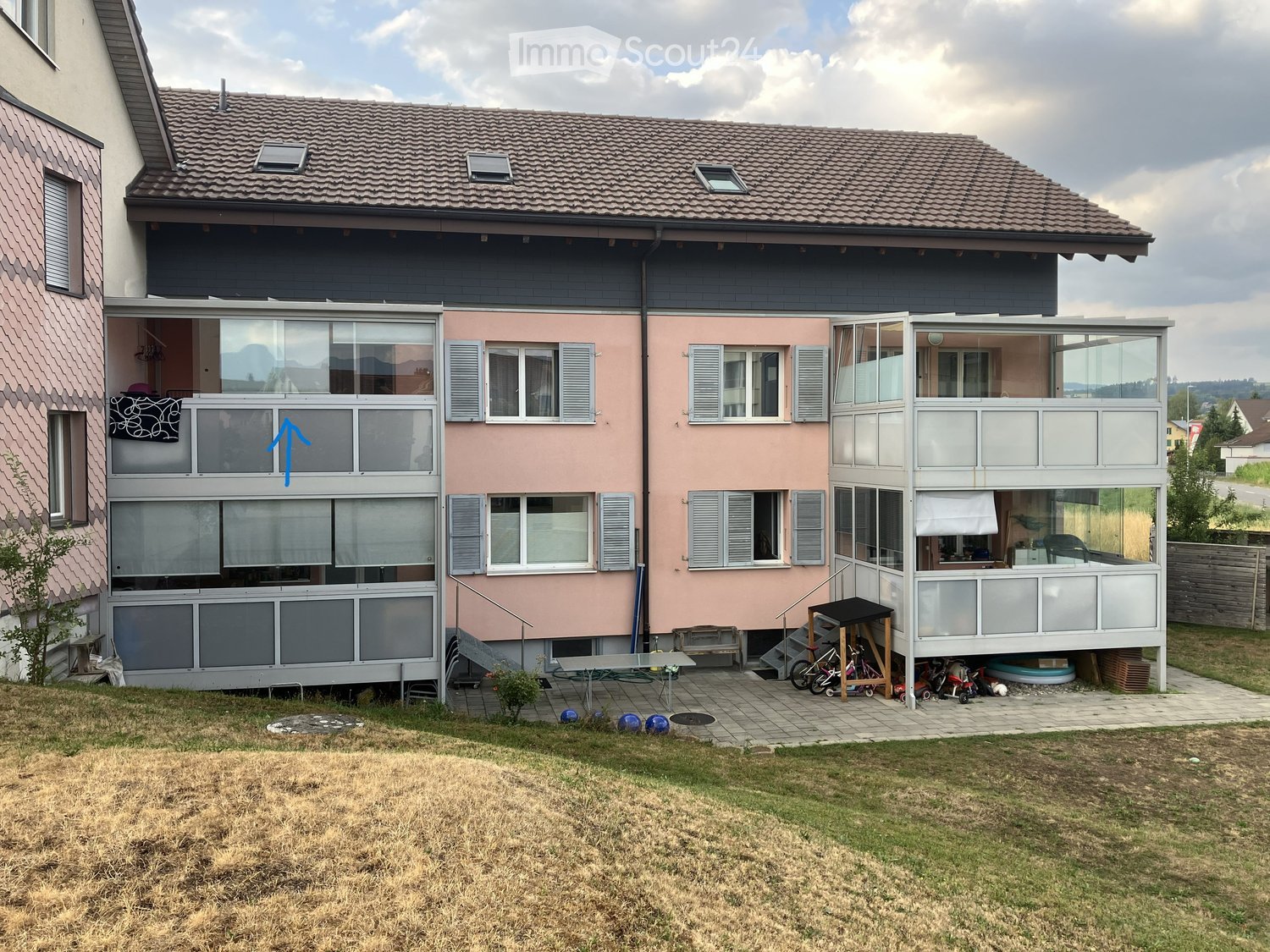 Two-story house with pink and grey exterior, multiple windows, balconies, and a covered patio area. The yard includes a small table, chairs, and bicycles.