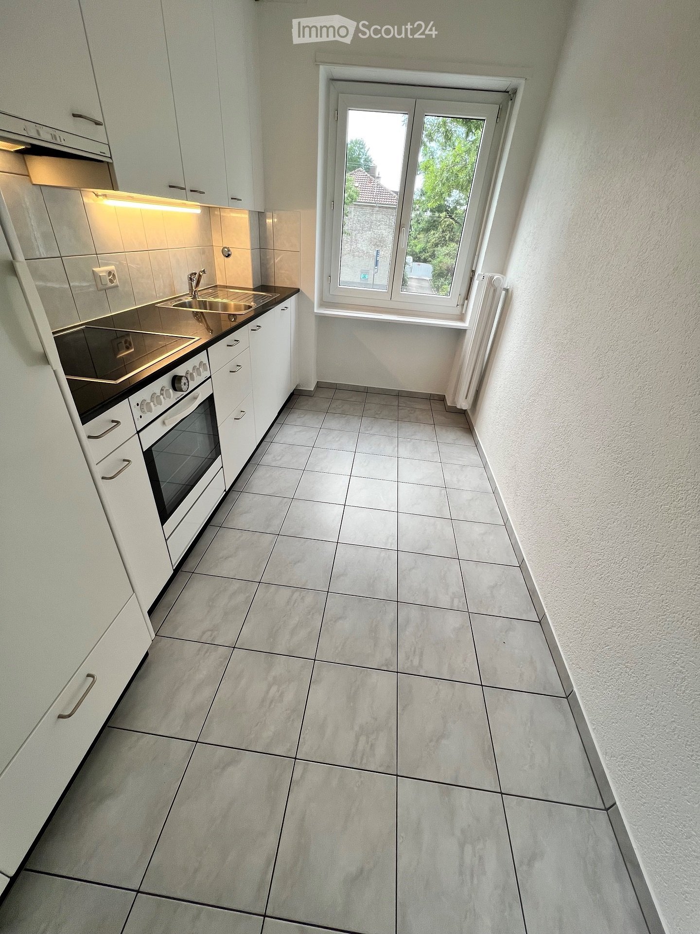 Modern kitchen with white cabinets, tiled floor, built-in oven, sink, and window overlooking greenery.