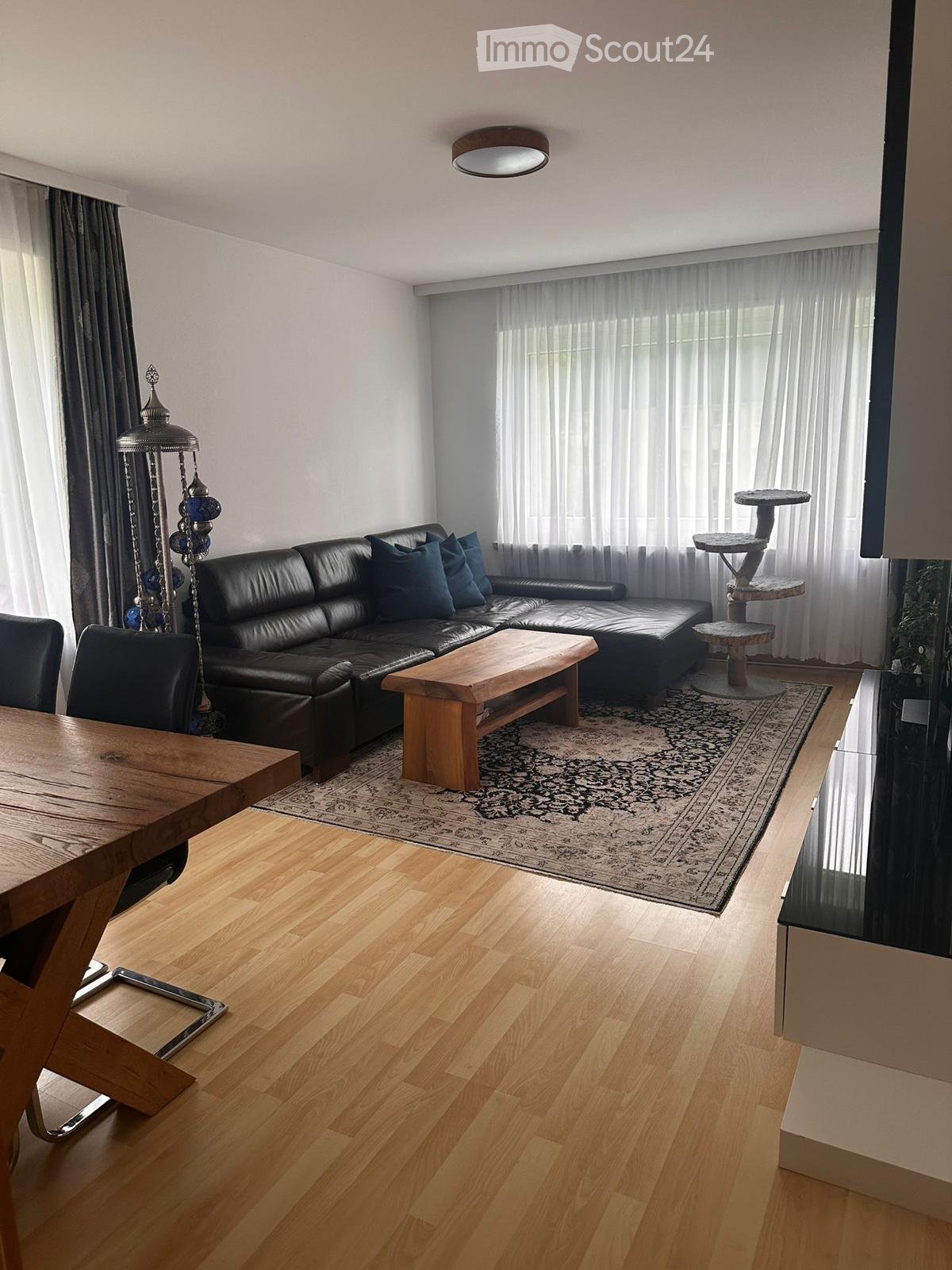 Modern living room with a black leather sofa, wooden coffee table, dining table, chairs, and a cat tree. The room features large windows with sheer curtains, a patterned rug, and a decorative floor lamp.