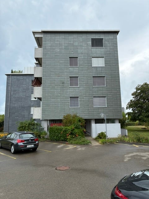 Modern multi-story apartment building with grey facade, balconies, and a parking area in front.