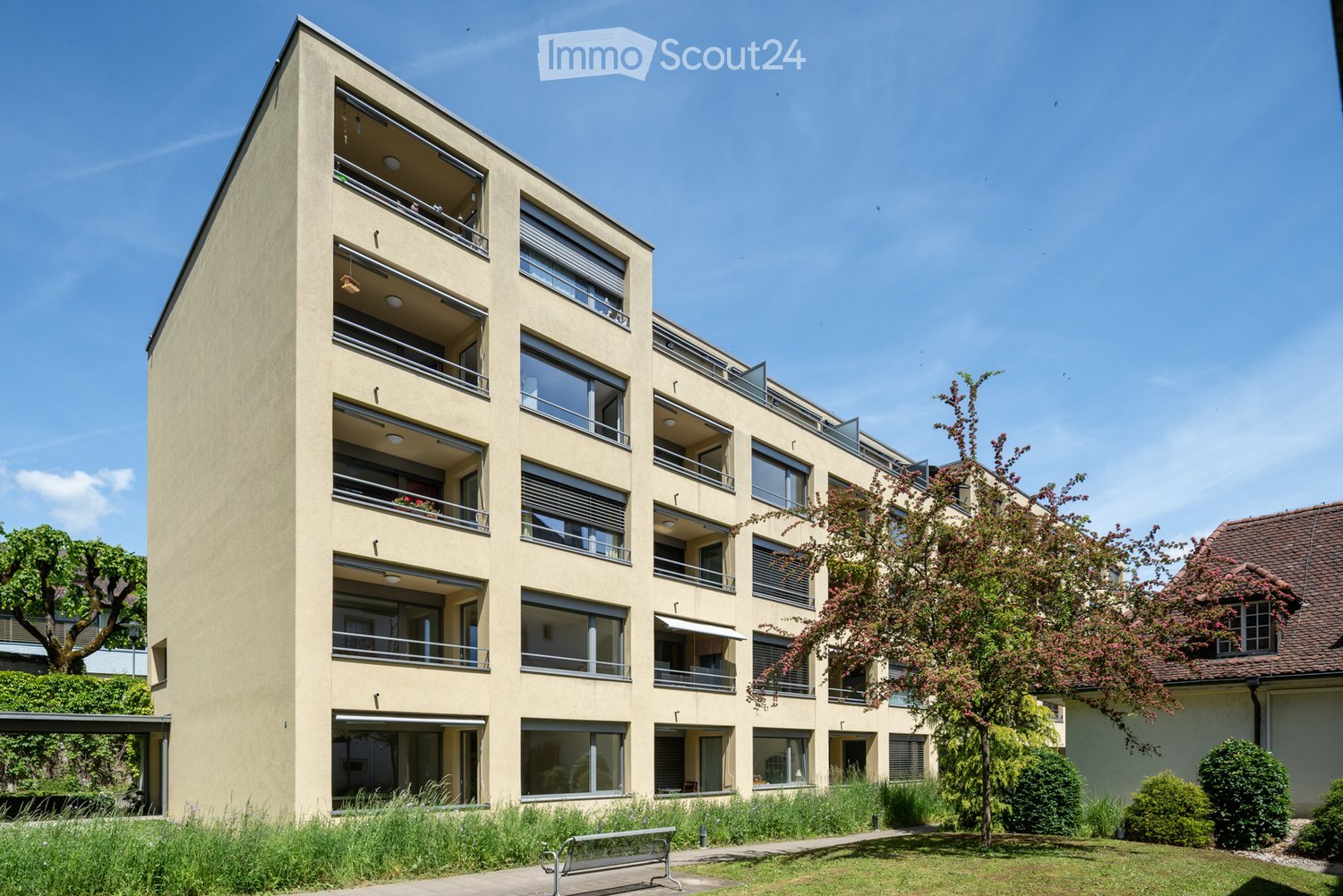 Modern multi-story residential building with balconies, large windows, and a landscaped garden area in front.