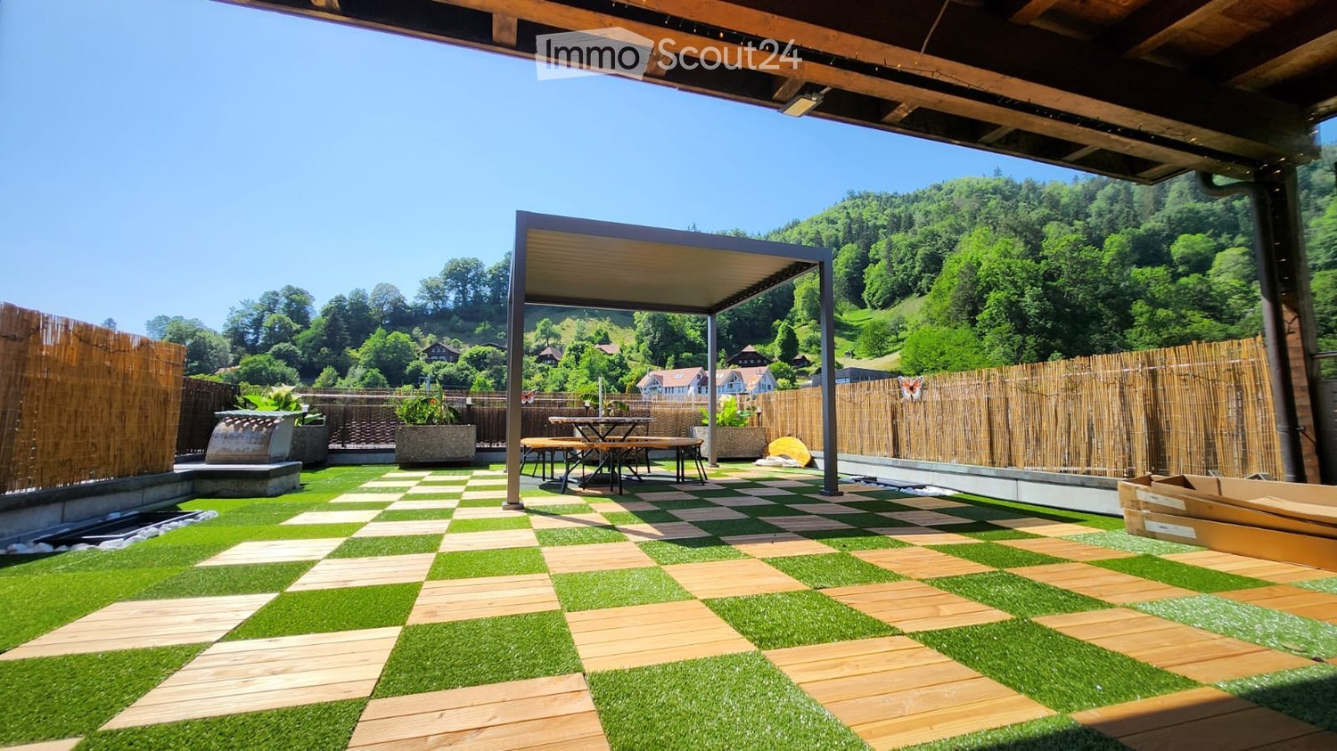 Outdoor patio with artificial grass and wooden tiles, a covered seating area with a table and benches, surrounded by bamboo fencing and potted plants, overlooking a scenic valley.