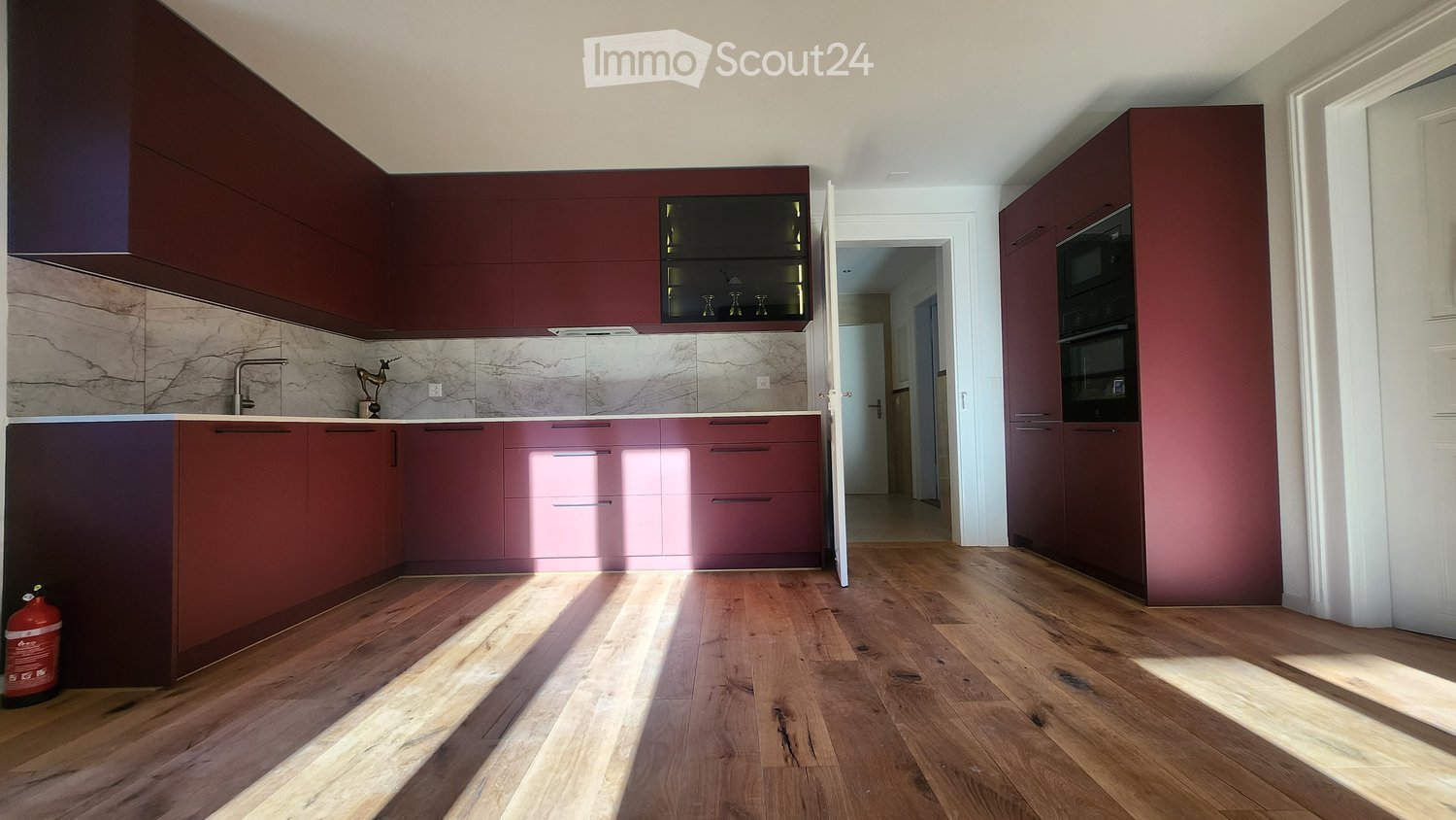 Modern kitchen with dark red cabinets, marble backsplash, wooden flooring, and built-in appliances including an oven and fridge.