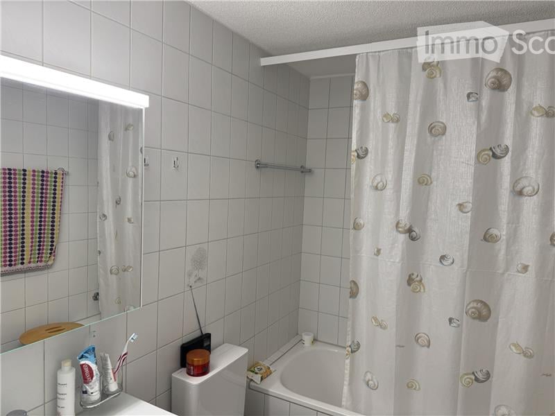 A small bathroom with white tiled walls and floor, featuring a bathtub with a shell-patterned shower curtain, a toilet, a mirror with a light fixture above it, and a towel hanging on the wall. Personal care items are placed on the sink counter.