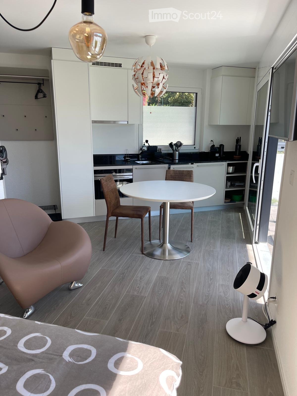 open plan kitchen with dining area, modern style, large dining table with 4 chairs, stainless steel kitchen island with sink, white cabinets, modern pendant light, wooden floor, open door, white sheet on bed