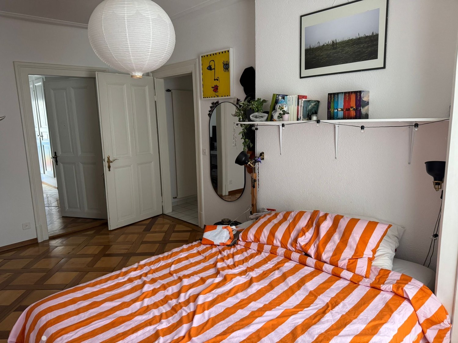 Cozy bedroom with a wooden floor, a bed with orange and white striped bedding, a mirror, a bookshelf with books and plants, and a hanging paper lantern.