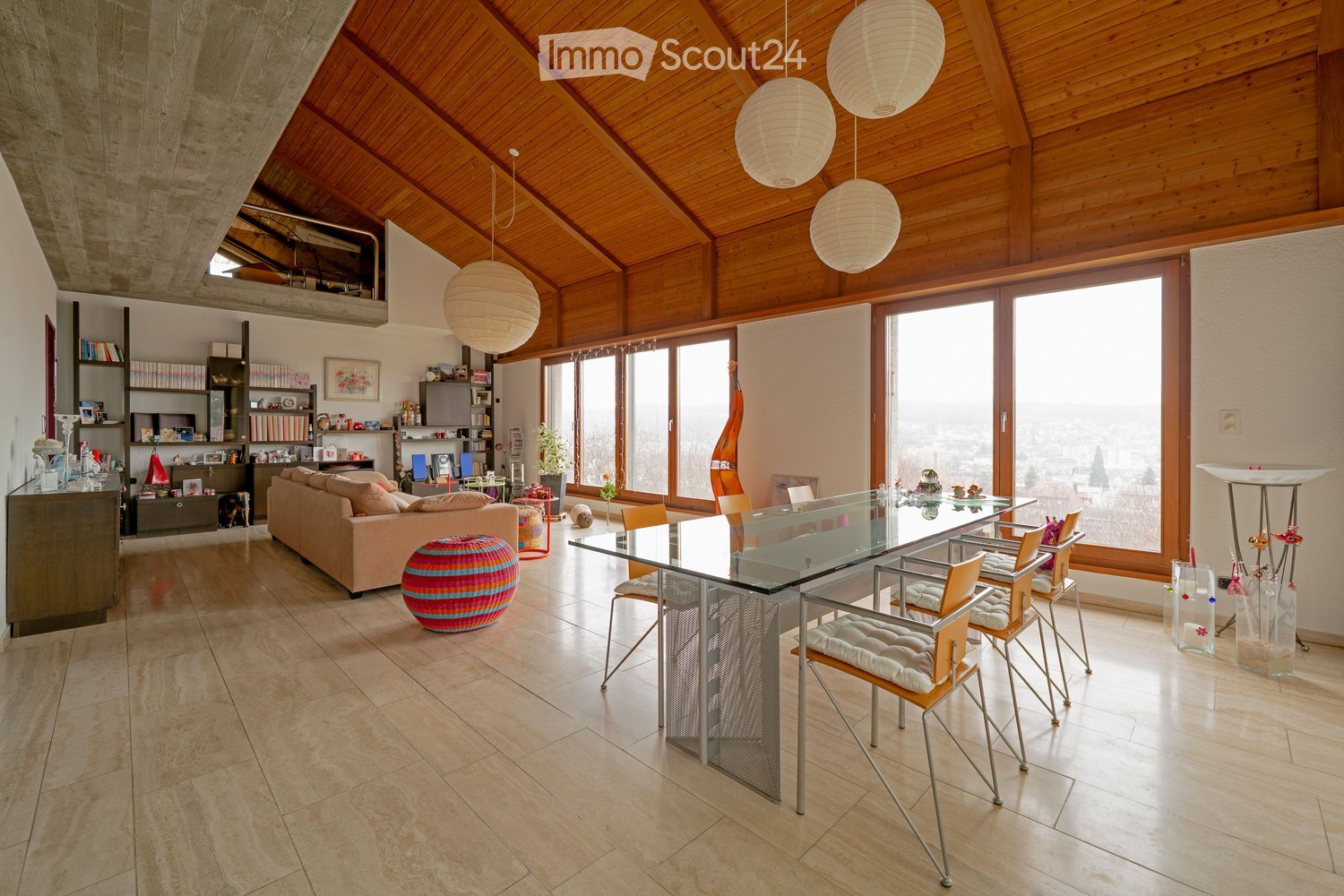 A spacious living room with a high, wooden-beamed ceiling, large windows offering a view, a beige sofa, a colorful ball, a glass dining table with chairs, shelves with books and decorative items, and pendant lamps.