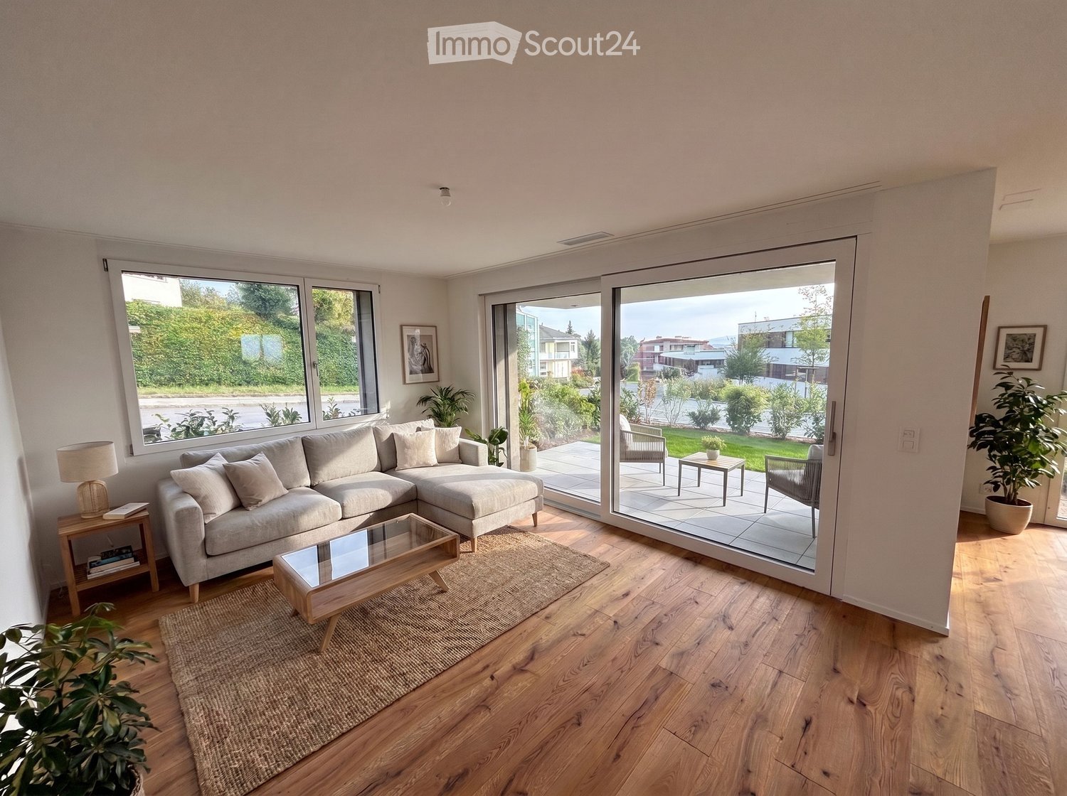 Modern living room with light wooden flooring, large windows, and a sliding glass door leading to a patio. Features a grey sectional sofa, coffee table, side table, floor lamp, and potted plants.