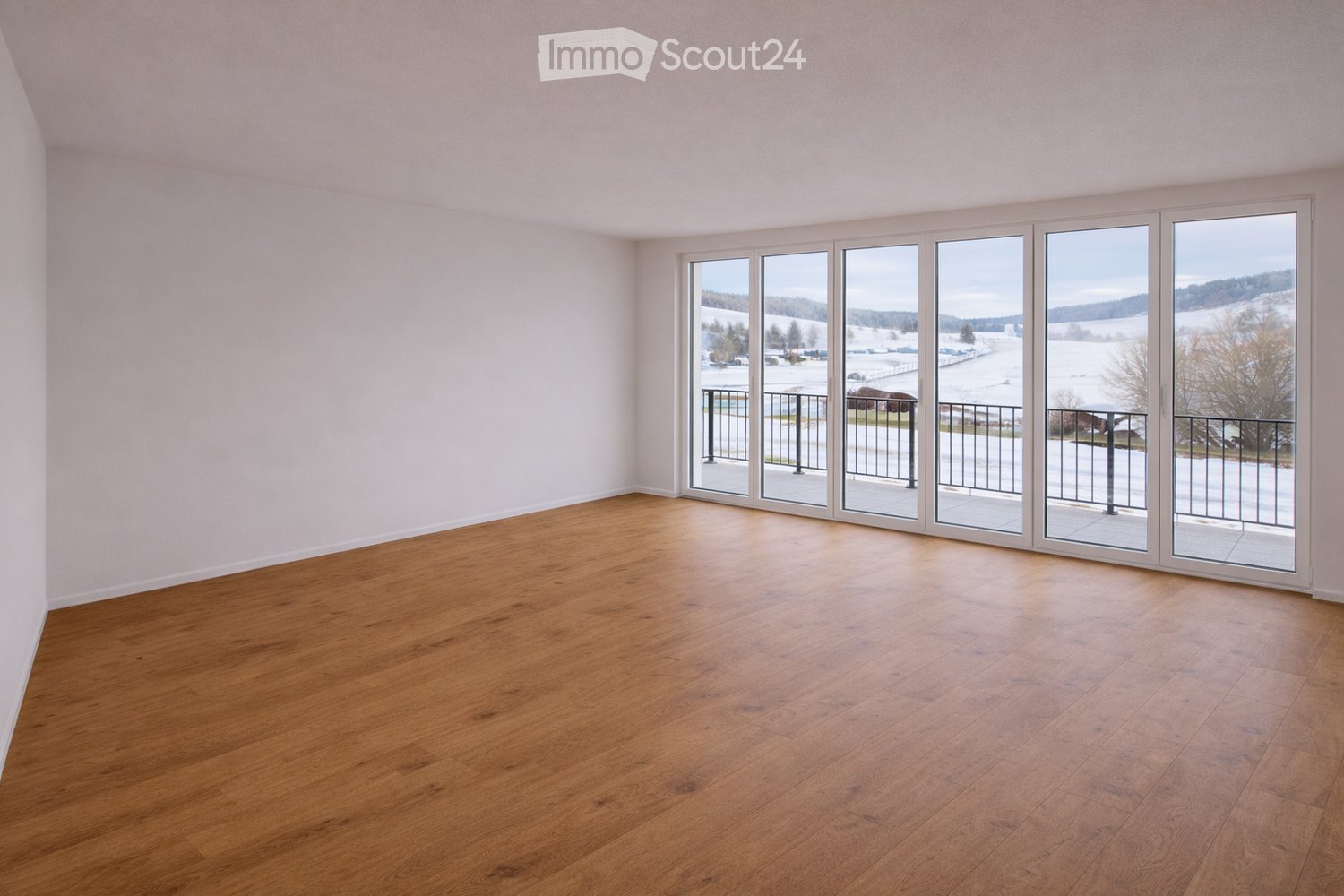 Empty room with wooden flooring and large sliding glass doors leading to a balcony with a view of a snowy landscape.