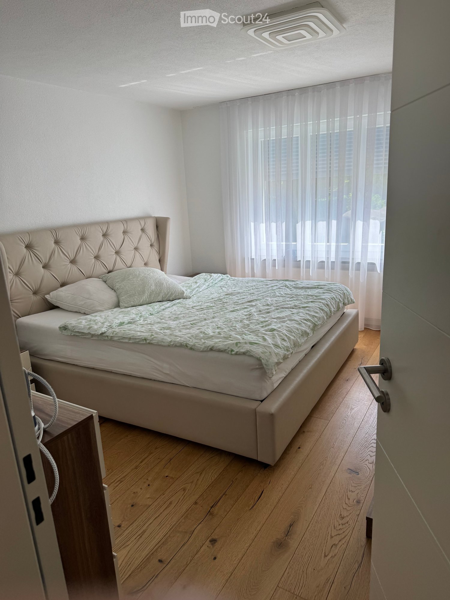Modern bedroom with a tufted headboard bed, light green bedding, wooden floor, and large window with sheer curtains.