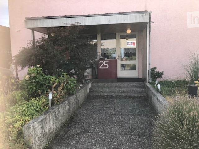 Entrance to a building with a small staircase leading to a double door, number 25 displayed on a red sign, surrounded by low shrubs and plants.