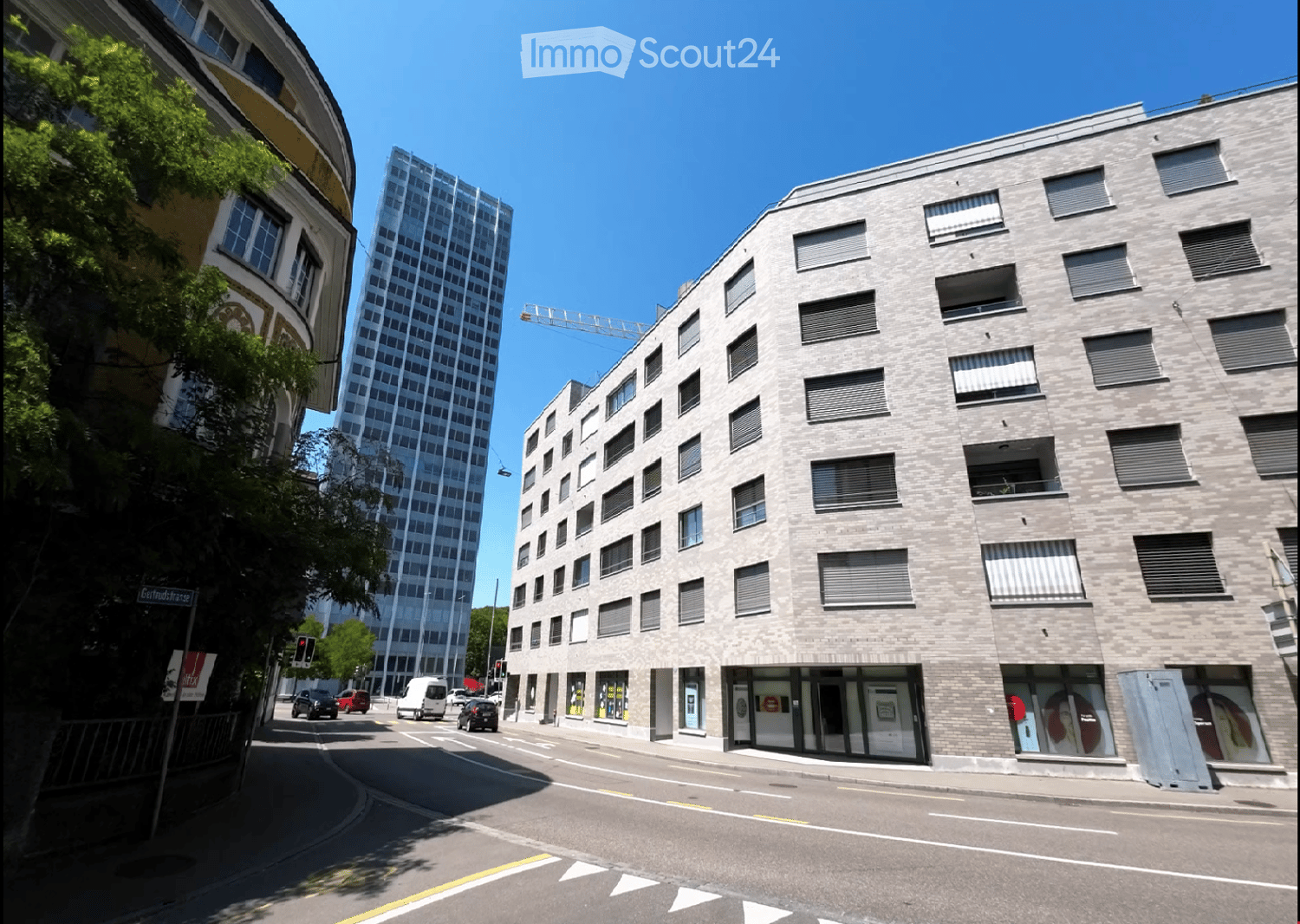 An exterior shot of an apartment building. Located on a city street with road signs, cars, trees, and street lights. The building has multiple floors with windows and shutters. There is a crane at the top of the building behind it.