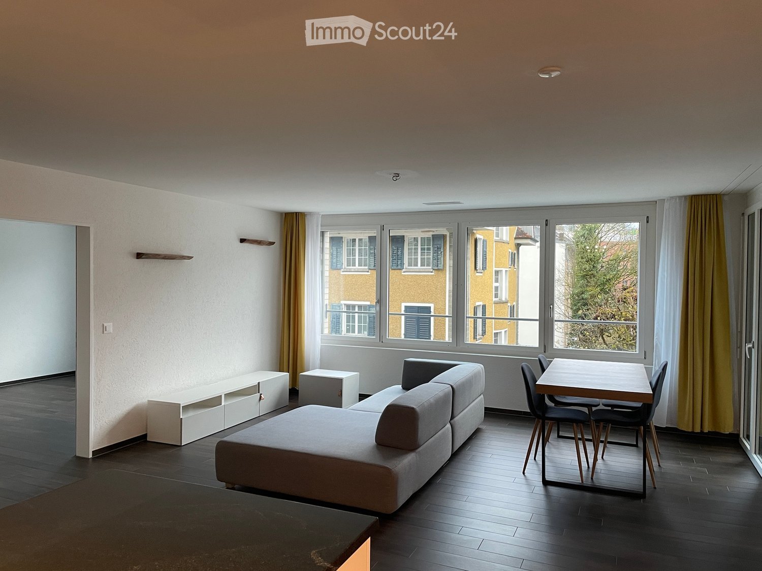 Modern living room with dark wooden flooring, large windows, grey sectional sofa, small table with chairs, and white shelving unit.