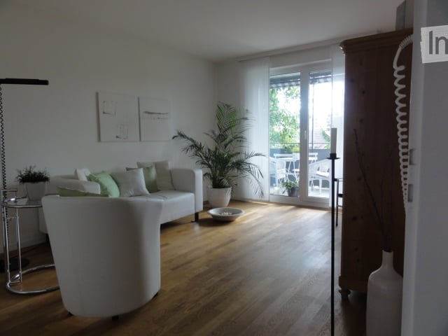 Bright living room with white walls, wooden floor, large windows, and a balcony. Features a white sofa with green and white cushions, a coffee table, an armchair, a floor lamp, a small table with a plant, and a large potted plant near the balcony.