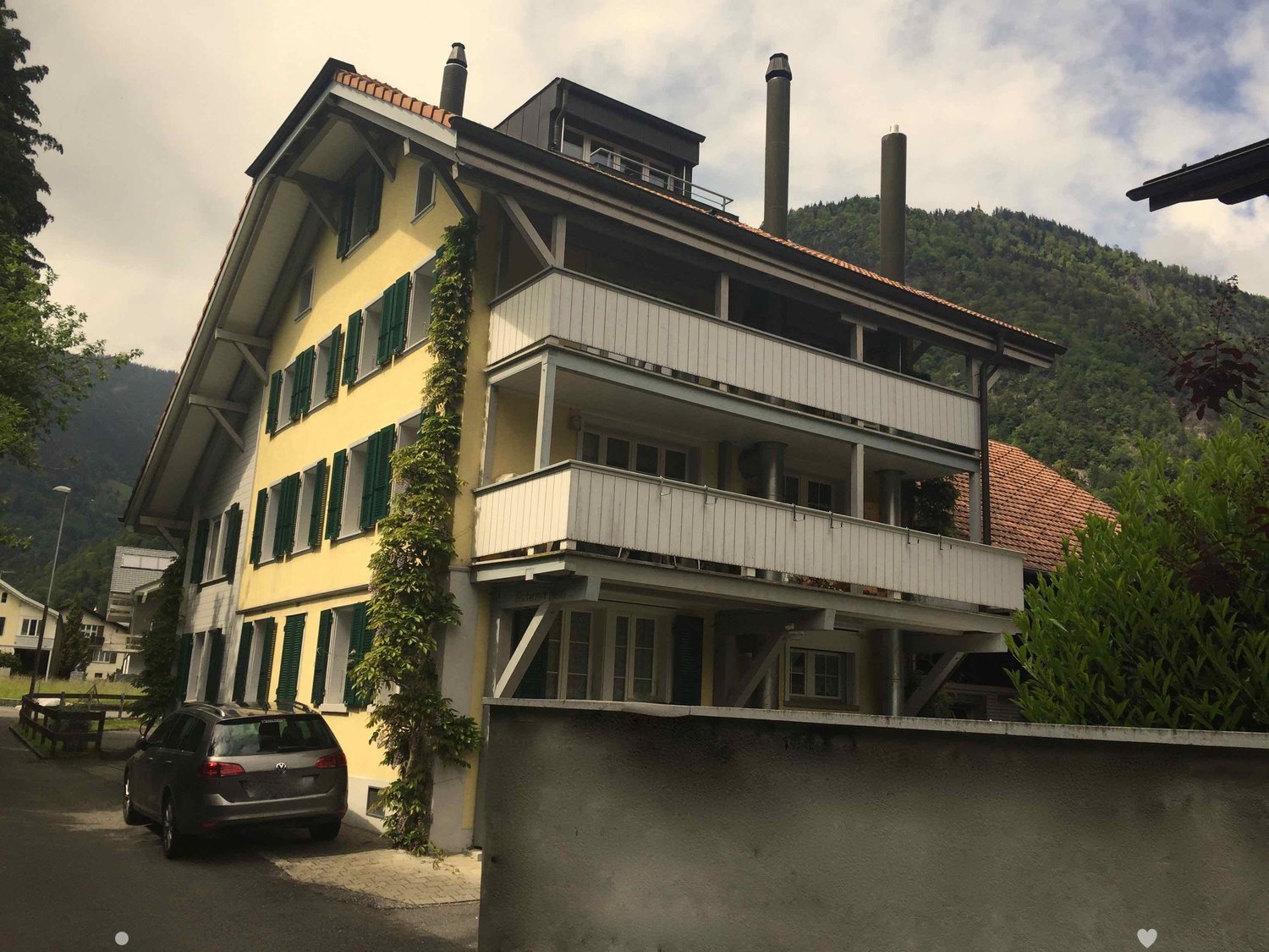 3-story yellow house, green shutters, brick chimneys, balcony on second floor, car parked in front