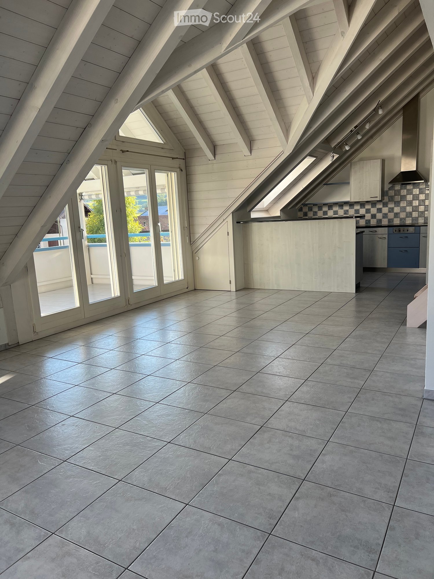 Bright, airy attic space with wooden beams, large skylights, and a modern kitchen area featuring light-colored cabinets and a dishwasher. The floor is tiled, and there is a sliding glass door leading to an outdoor area.