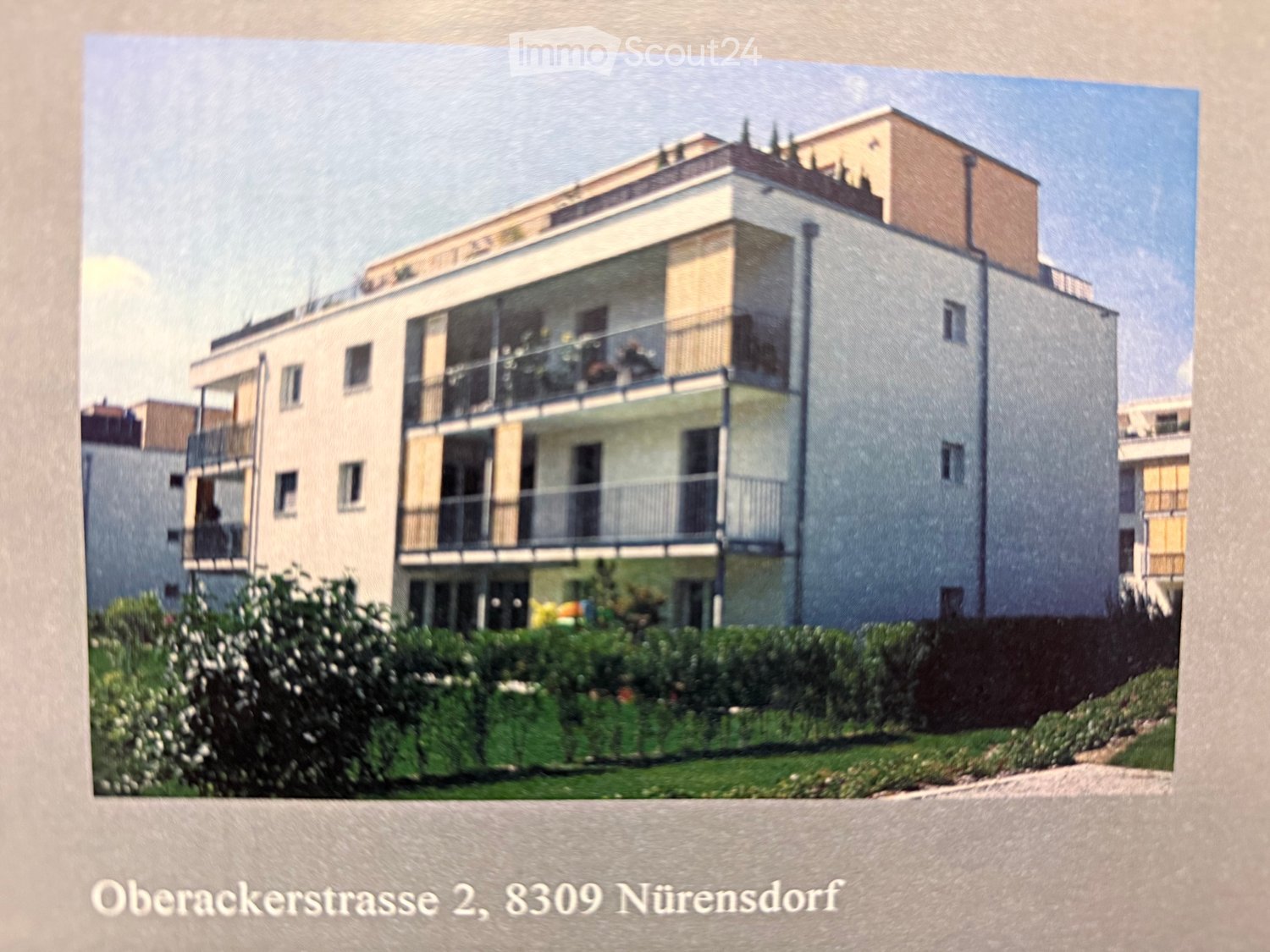 Modern multi-story residential building with balconies, surrounded by greenery and shrubs.