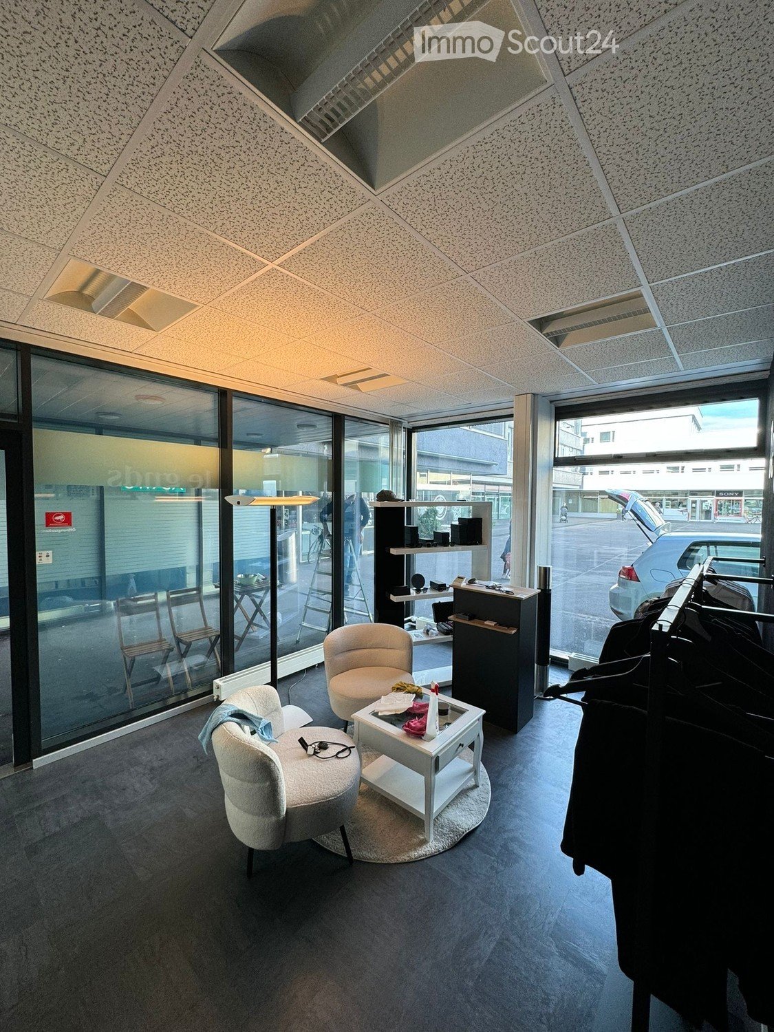 Modern, well-lit room with large windows, featuring two chairs, a small table, a clothing rack, and a shelf with various items.